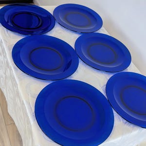 May include: Six cobalt blue glass plates are arranged on a white tablecloth. Each plate has a dark blue ring around the center. The plates are oval-shaped and appear to be new.