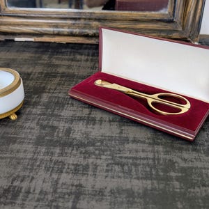 May include: A gold-colored pair of scissors in a red velvet-lined box, next to a white and gold ashtray. The scissors are open, and the ashtray has a gold-colored rest. The items are on a dark gray surface.