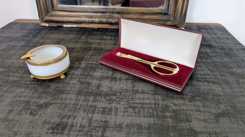 May include: A gold-colored pair of scissors in a red velvet-lined box, next to a white and gold ashtray. The scissors are open, and the ashtray has a gold-colored rest. The items are on a dark gray surface.
