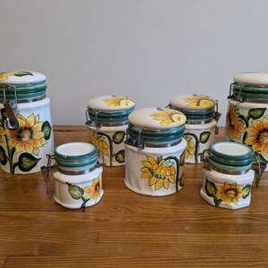 Peut inclure: Ensemble de six pots en céramique avec des motifs de tournesols. Les pots ont des couvercles verts avec un loquet en métal.