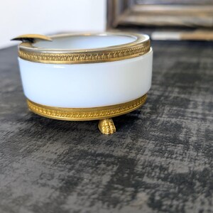May include: A white and gold ashtray with a cylindrical shape. The ashtray has a white body with gold trim at the top and bottom. The ashtray rests on three small, gold-colored feet. A small, gold-colored rest is on the side.