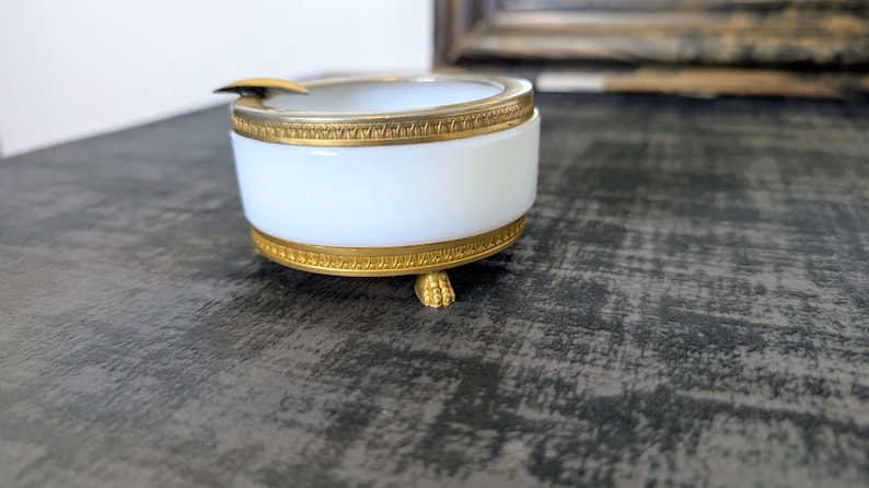 May include: A white and gold ashtray with a cylindrical shape. The ashtray has a white body with gold trim at the top and bottom. The ashtray rests on three small, gold-colored feet. A small, gold-colored rest is on the side.