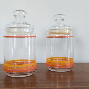 Puede incluir: Dos tarros de vidrio con diseños de rayas naranjas y amarillas. Los tarros tienen tapas y están vacíos.