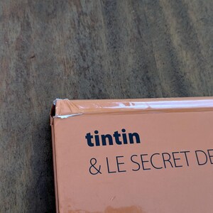 Puede incluir: Una cubierta de libro de color naranja claro con el texto negro "tintin & LE SECRET DE LA LICORNE". La esquina superior izquierda de la cubierta muestra un desgaste visible con un peque&ntilde;o desgarro en el papel y un ligero pliegue en el borde.