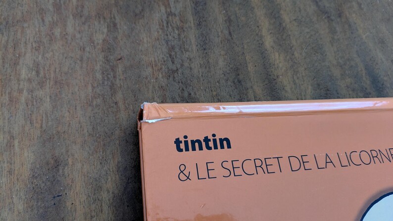 Puede incluir: Una cubierta de libro de color naranja claro con el texto negro "tintin & LE SECRET DE LA LICORNE". La esquina superior izquierda de la cubierta muestra un desgaste visible con un peque&ntilde;o desgarro en el papel y un ligero pliegue en el borde.