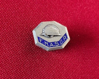 May include: A silver-toned octagonal lapel pin featuring an embossed helmet graphic above a blue enamel banner with the white text F.N.A.S.O.R. The pin has a metallic finish and a geometric shape, displaying clear lettering and a detailed helmet icon.