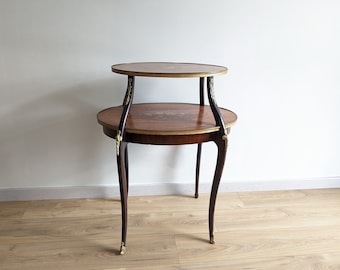 Puede incluir: Una mesa auxiliar redonda de dos niveles de madera con un diseño de marquetería. La mesa tiene un acabado marrón oscuro y detalles dorados. La mesa está sobre un suelo de madera clara.