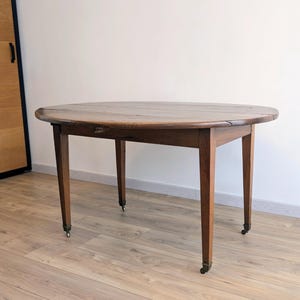 Peut inclure: Une table ronde en bois avec quatre pieds et de petites roulettes. La table est de couleur marron et semble être ancienne. La table est placée contre un mur blanc et une porte en bois.