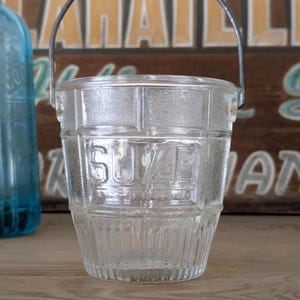 Peut inclure: Un seau en verre transparent avec une poignée en métal. Le mot "SUZE" est gravé sur le devant. Le seau a un design côtelé et repose sur une surface en bois. Une bouteille en verre bleu est visible en arrière-plan.