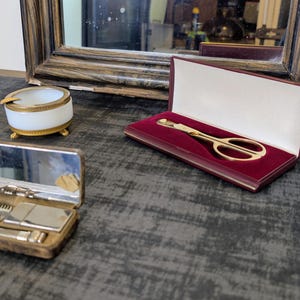 May include: An antique vanity set with a mirror, a small ashtray, and a gold-toned scissor set in a burgundy case. The set also includes a small, open, brown case with a mirror and grooming tools.