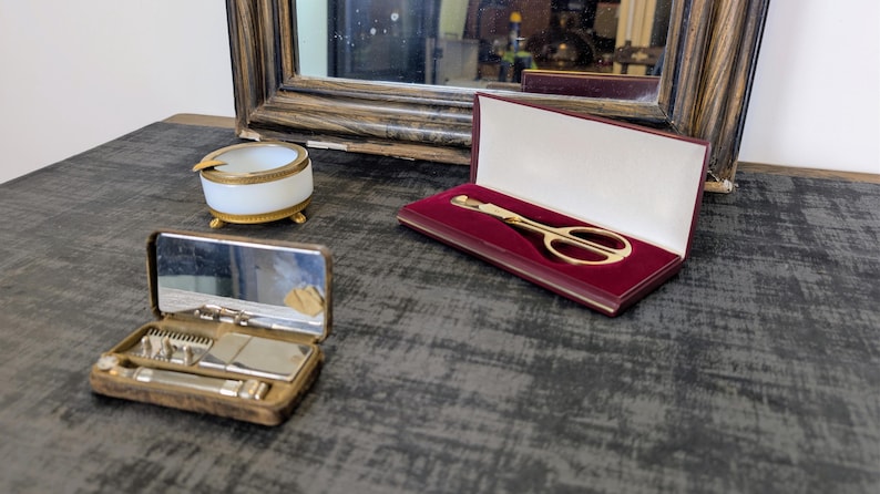 May include: An antique vanity set with a mirror, a small ashtray, and a gold-toned scissor set in a burgundy case. The set also includes a small, open, brown case with a mirror and grooming tools.