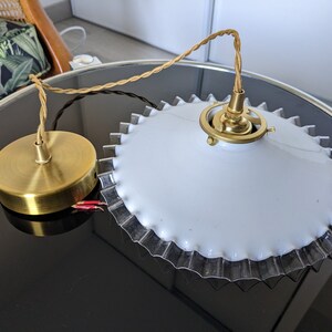 May include: A white ceiling light fixture with a gold metal base and a scalloped edge. The light fixture is hanging from a brown cord.