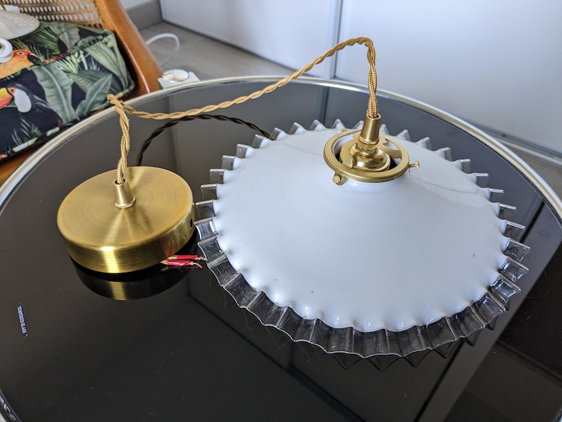 May include: A white ceiling light fixture with a gold metal base and a scalloped edge. The light fixture is hanging from a brown cord.