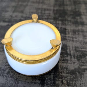 May include: A white and gold ashtray with a circular design. The ashtray has a white interior and a gold rim, with three gold rests for cigarettes. The ashtray is a decorative item.