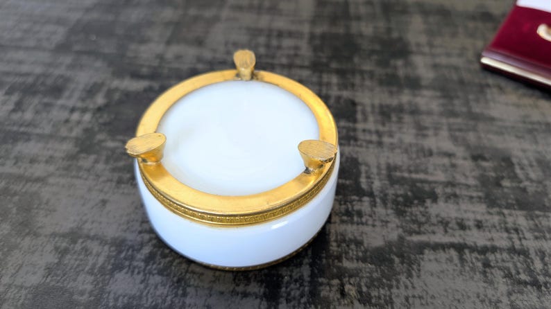 May include: A white and gold ashtray with a circular design. The ashtray has a white interior and a gold rim, with three gold rests for cigarettes. The ashtray is a decorative item.
