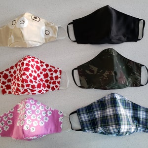 Op de afbeelding: Zes verschillende stoffen mondmaskers met verschillende patronen. De maskers zijn allemaal van katoen en hebben elastische oorlussen. De patronen omvatten een beige masker met bruine berengezichten, een rood masker met witte esdoornbladeren, een roze masker met witte bloemen, een zwart masker, een camouflagemasker en een blauw-wit geruit masker.