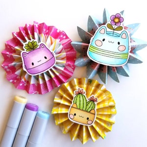 May include: Three paper fan crafts with cute cat and cactus illustrations. Each fan is decorated with a different patterned paper. The cat illustrations are in pink, purple, and blue. The cactus illustration is in green and yellow.