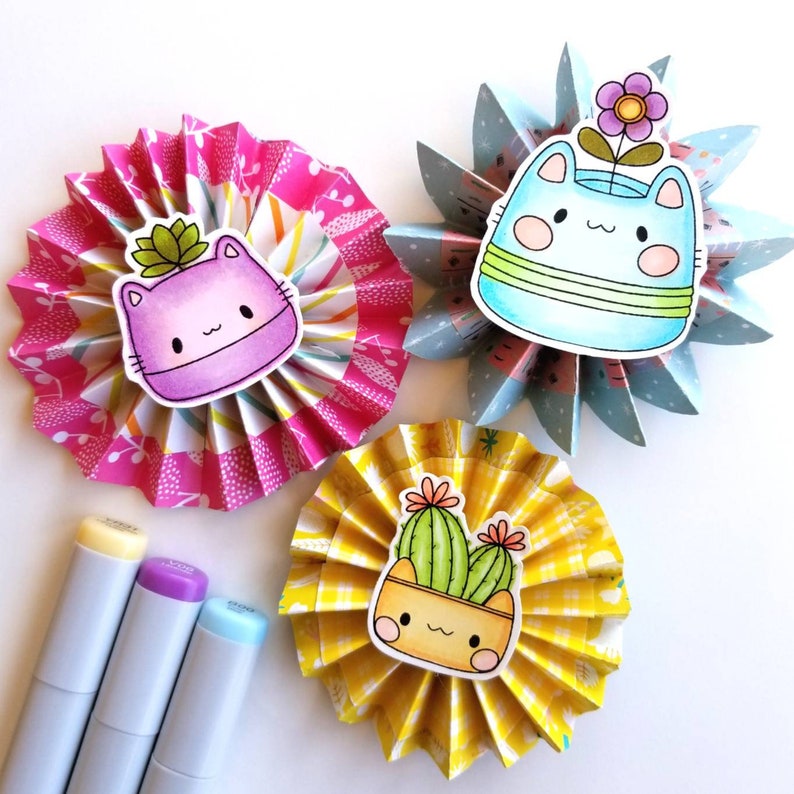 May include: Three paper fan crafts with cute cat and cactus illustrations. Each fan is decorated with a different patterned paper. The cat illustrations are in pink, purple, and blue. The cactus illustration is in green and yellow.