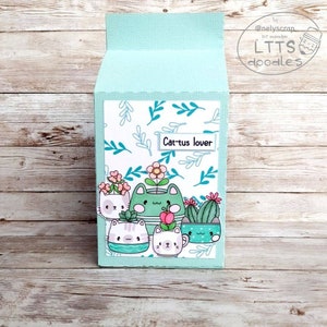 May include: A light blue card with a white background featuring a pattern of green leaves. The card has a white label with the text "Cactus lover" and an illustration of five cats in flower pots.