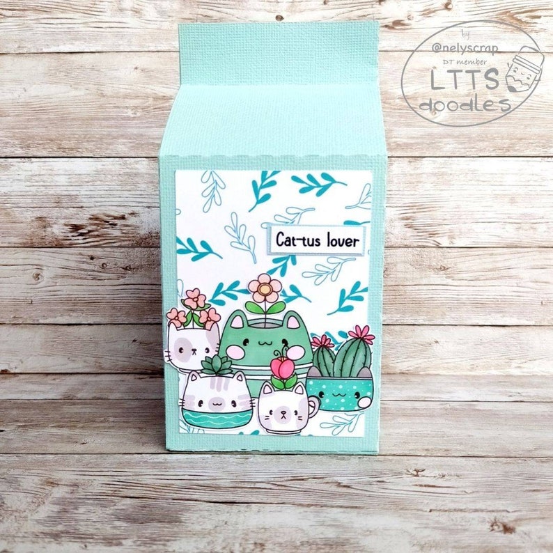 May include: A light blue card with a white background featuring a pattern of green leaves. The card has a white label with the text "Cactus lover" and an illustration of five cats in flower pots.