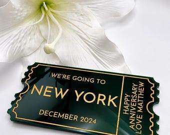Può includere: Una targa in acrilico verde scuro a forma di biglietto con scritte dorate che recitano WE'RE GOING TO NEW YORK, DECEMBER 2024 e HAPPY ANNIVERSARY LOVE MATTHEW. La targa è posizionata davanti a un grande fiore di giglio bianco.