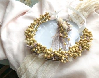 May include: A dried flower crown with a white ribbon, featuring small yellow and purple flowers. The crown is displayed on a white plate with a small bouquet of dried flowers.