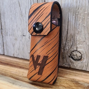 May include: A brown leather sheath with black tiger stripe pattern and a black snap closure. The sheath has the initials "VH" embossed on the front. The background is a weathered wooden surface.