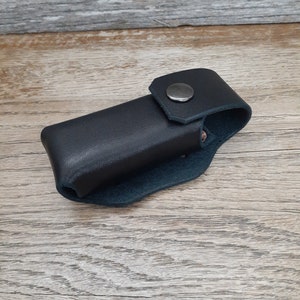May include: A black leather case with a snap closure. The case is designed to hold a small object, such as a knife or a multi-tool.