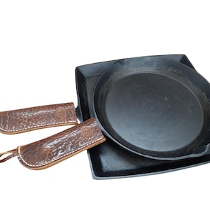 May include: A black cast iron round skillet and a black cast iron square griddle. Both pans feature brown textured leather handle covers with white stitching and small loops at the ends. The leather covers are embossed with a circular logo.