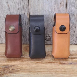 Pode incluir: Três bainhas de couro em marrom, preto e bege, cada uma com fecho de pressão. As bainhas são retangulares com uma presilha na parte superior. A bainha marrom tem um fecho de pressão de latão, a preta tem um fecho de pressão preto e a bege tem um fecho de pressão preto.