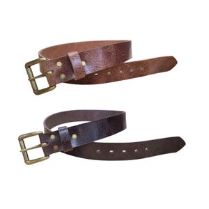 Puede incluir: Dos cinturones de cuero marrón, cada uno con una hebilla rectangular de latón y remaches a juego. Los cinturones están enrollados, mostrando su textura y diseño. El cinturón superior es de color marrón claro, el cinturón inferior es de color marrón oscuro.
