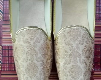 May include: A pair of gold and beige patterned slippers with a gold trim. The slippers are on a red and gold patterned surface.