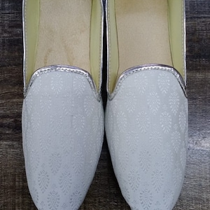 May include: A pair of white, silver-trimmed, flat shoes with a floral pattern. The shoes have a pointed toe and a slip-on design. D.No.1008