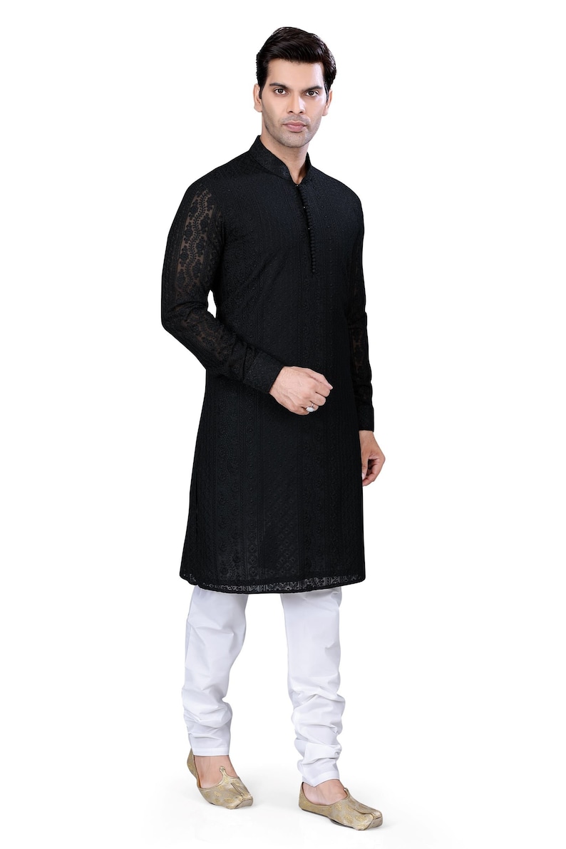 May include: A black long-sleeved kurta with intricate embroidery. The kurta has a mandarin collar and a button closure. The model is wearing white pants and gold shoes.