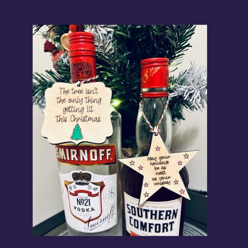 Peut inclure: Deux bouteilles de liqueur miniatures avec des ornements en bois sur le th&egrave;me de No&euml;l. La bouteille de vodka Smirnoff a un ornement de sapin de No&euml;l vert avec le texte "The tree isn't the only thing getting lit this Christmas". La bouteille de Southern Comfort a un ornement en forme d'&eacute;toile avec le texte "May your holidays be as neat as your whiskey".