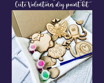 Puede incluir: Un kit de pintura DIY con recortes de madera de una mariposa, un pulpo, un caracol, una tortuga y un oso sosteniendo un corazón. El kit incluye cuatro pequeños frascos de pintura y un pincel. El kit está empaquetado en una caja de cartón marrón.