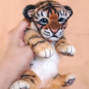 Peut inclure: Une peluche artisanale de bébé tigre avec des rayures orange et noires, des yeux bleus et un ventre blanc. Le bébé tigre a un visage et des pattes détaillés, et est tenu par une main. La peluche est une représentation mignonne et câline d'un bébé tigre.