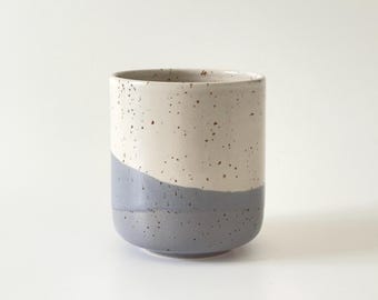 Może przedstawiać: Cylindryczny kubek ceramiczny o dwukolorowym wzorze, z nakrapianą kremową górną częścią i jednolitym szarym dołem. Kubek posiada gładką, zaokrągloną podstawę oraz prostą krawędź, prezentując wyraźny podział kolorystyczny na całej powierzchni.