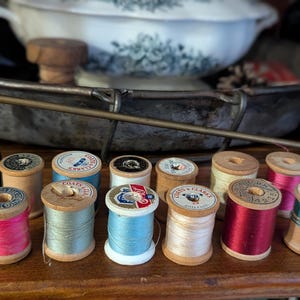 May include: A collection of twelve vintage wooden spools of thread in various colors including bright pink, light blue, white, cream, and deep red. Each spool features a circular paper label on top with brand text like Coats and Clark.