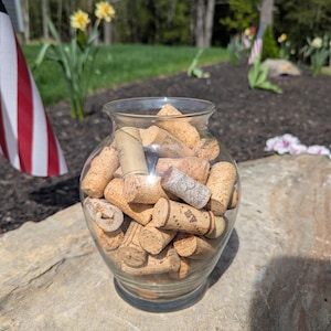 May include: A clear glass vase filled with numerous corks. The corks are various shades of brown and beige, with some displaying printed text. The vase sits on a large, gray rock, with a blurred outdoor background.