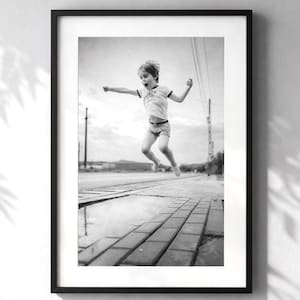 Könnte beinhalten: Schwarz-weiß gerahmtes Foto eines Kindes, das über eine Pfütze auf einer Ziegeloberfläche springt. Das Kind trägt ein T-Shirt und Shorts. Der Hintergrund zeigt Stromleitungen und einen bewölkten Himmel.