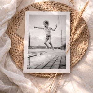 May include: Black and white photograph of a child jumping over a puddle. The image is set on a woven placemat with wheat stalks and a sheer white fabric. The child is wearing a t-shirt and shorts.