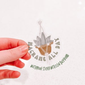 Puede incluir: Una pegatina transparente con una ilustración verde y marrón de una planta en maceta colgada de un macramé para plantas. El texto "Macrame All Day" está escrito alrededor de la planta en una fuente verde. La pegatina está etiquetada como "Pegatina impermeable con fondo transparente."