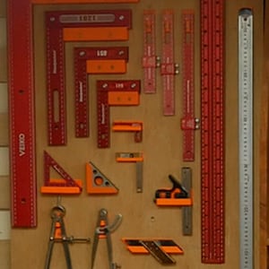 May include: A collection of red and orange woodworking tools, including rulers, squares, and a compass, are displayed on a wooden board. The tools are arranged in rows and columns, and some of them have labels with numbers on them.