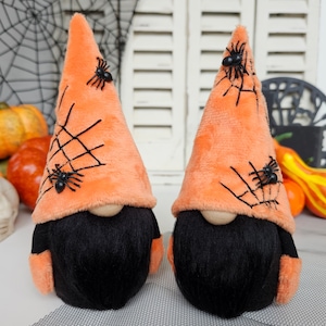 May include: Two orange Halloween gnomes with black beards and spider web designs on their hats. Each gnome has a black spider on their hat.