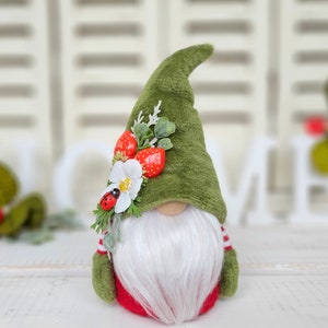 Può includere: Un gnomo di feltro verde con la barba bianca e una camicia a righe rosse e bianche. Il gnomo indossa un cappello di feltro verde con fragole rosse e un fiore bianco.