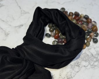 May include: A black, velvet-like headband lies on a marble surface. The headband is soft and appears to be made of a plush material. A collection of small, polished stones in various colours sits in the background.