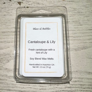 May include: A clear plastic clamshell package containing a soy blend wax melt. The label reads "Cantaloupe & Lily" with a description of fresh cantaloupe and lily. The net weight is 2.5 oz (70 g).
