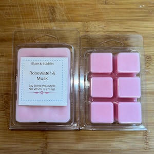 May include: Two clear plastic clamshell containers holding pink wax melts. One container has a solid block of wax with a label that reads "Blaze & Bubbles Rosewater & Musk Soy Blend Wax Melts Net Wt 2.5 oz (70.9g)." The other container has six square wax melts.