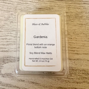May include: A clear plastic container holding a soy blend wax melt. The label reads "Gardenia" with a floral and orange scent description. The net weight is 2.5 oz (70 g). The brand name "Blaze & Bubbles" is also visible.